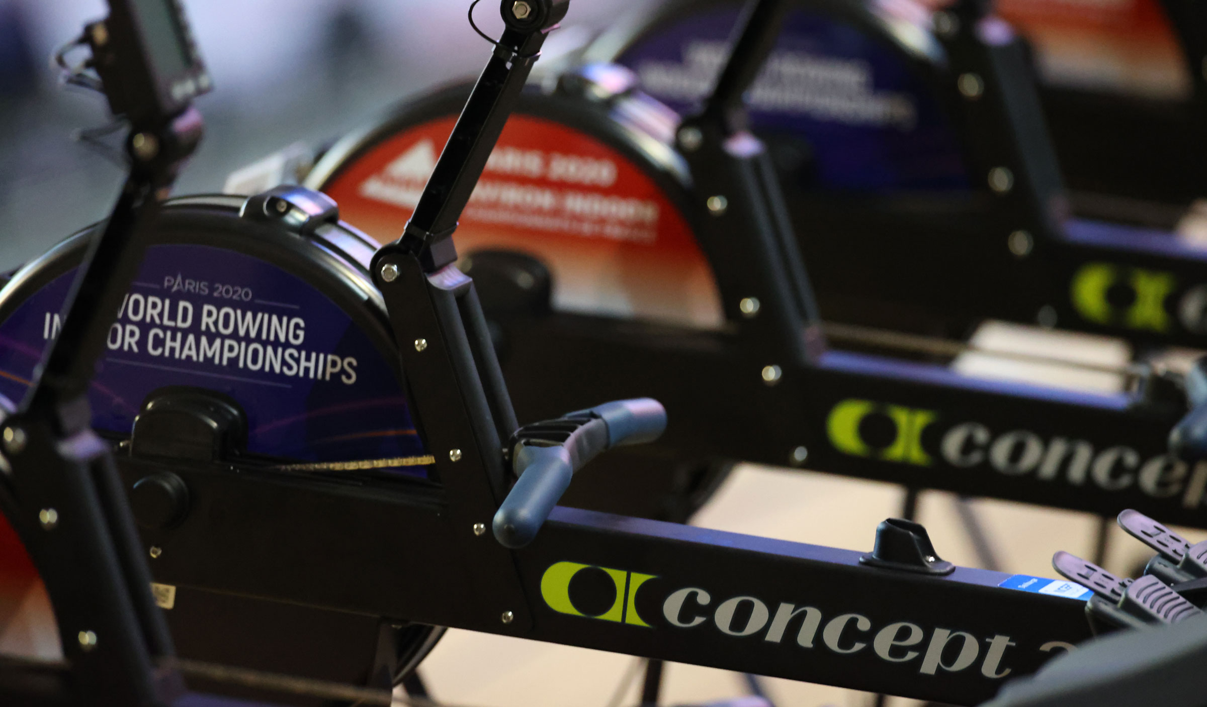 A close-up view of several indoor rowing machines lined up side by side. The machines are branded "Concept2", and one features a visible round panel labeled "Paris 2020 World Rowing Indoor Championships". The camera focuses on the flywheel and handle area, emphasizing the competitive, professional setup.