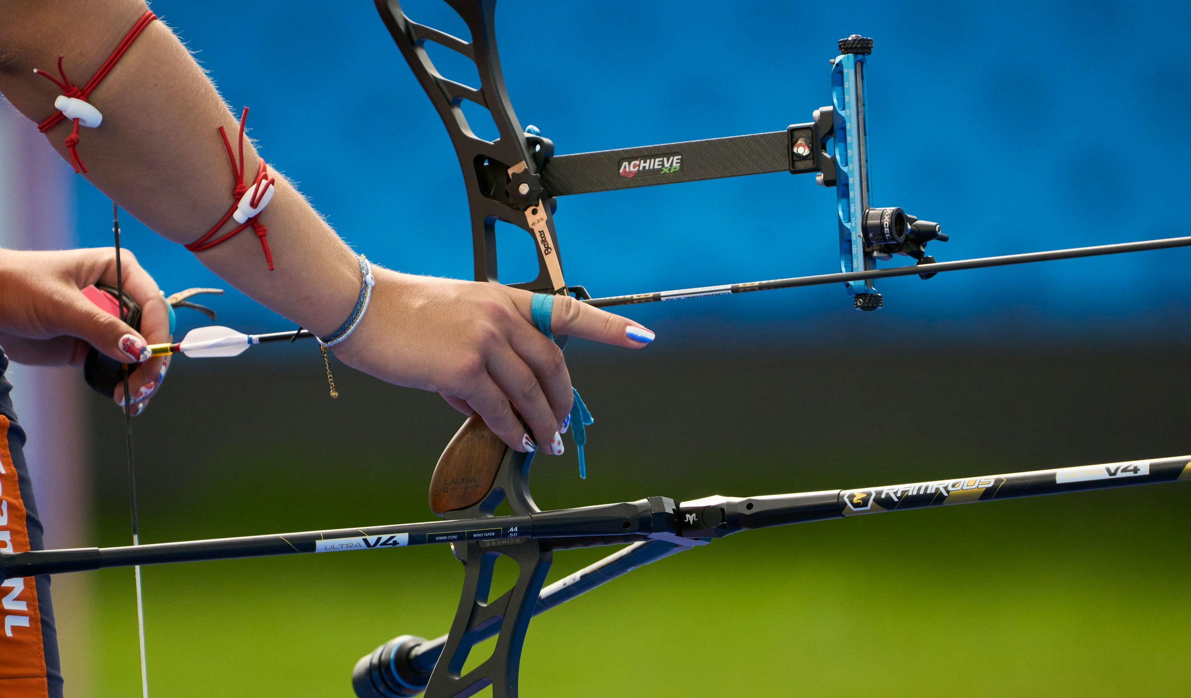 Close-up of a female archer’s hands preparing to shoot. The athlete grips a modern recurve bow with a nocked arrow and precision sight. Her nails are painted, and her arms are decorated with red cords and bracelets. The background is a blurred blue and green field.