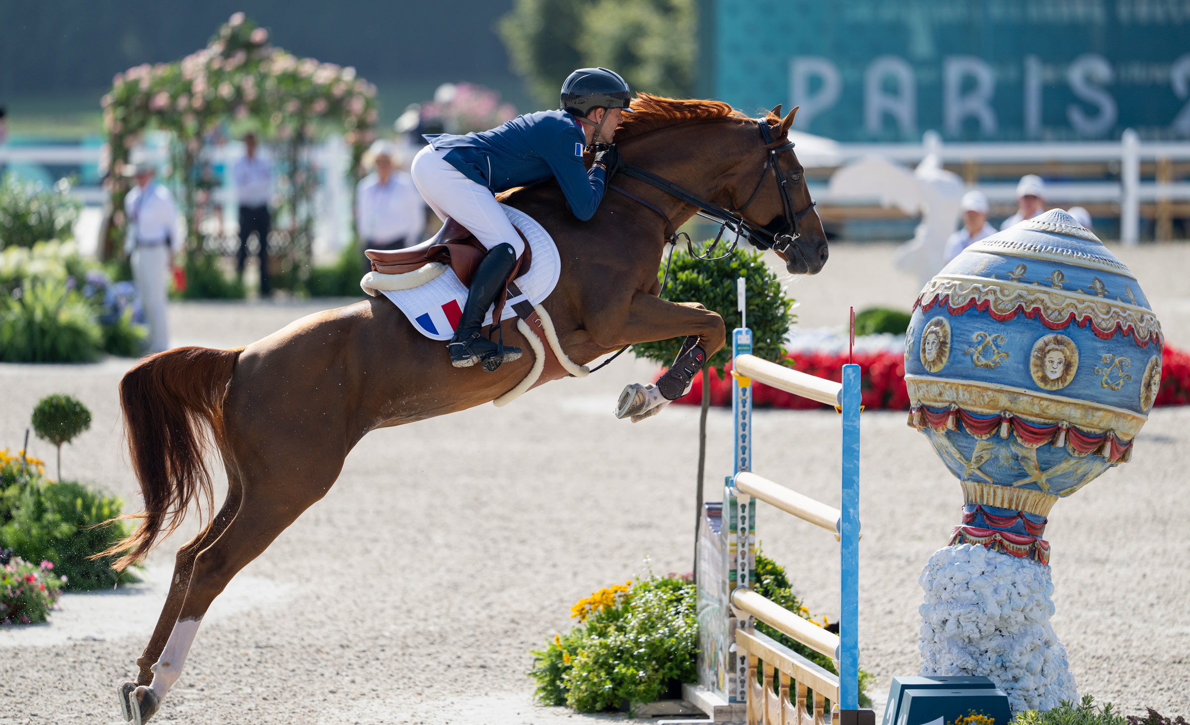 A French equestrian rider in blue and white uniform guides a chestnut horse mid-jump over a decorated obstacle during a show jumping event. The colorful jump includes a blue and gold orb with classical motifs. A blurred “PARIS 2024” sign and flower arrangements appear in the background.