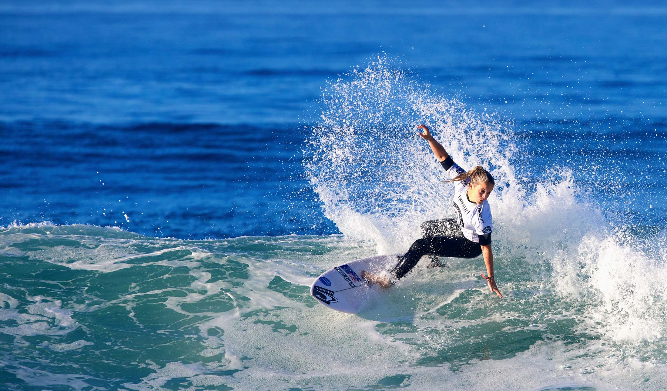 A female surfer carves across the face of a wave, sending a high spray of water into the air. She is wearing a white competition jersey and black wetsuit bottoms, balancing skillfully on her board against the deep blue ocean backdrop.