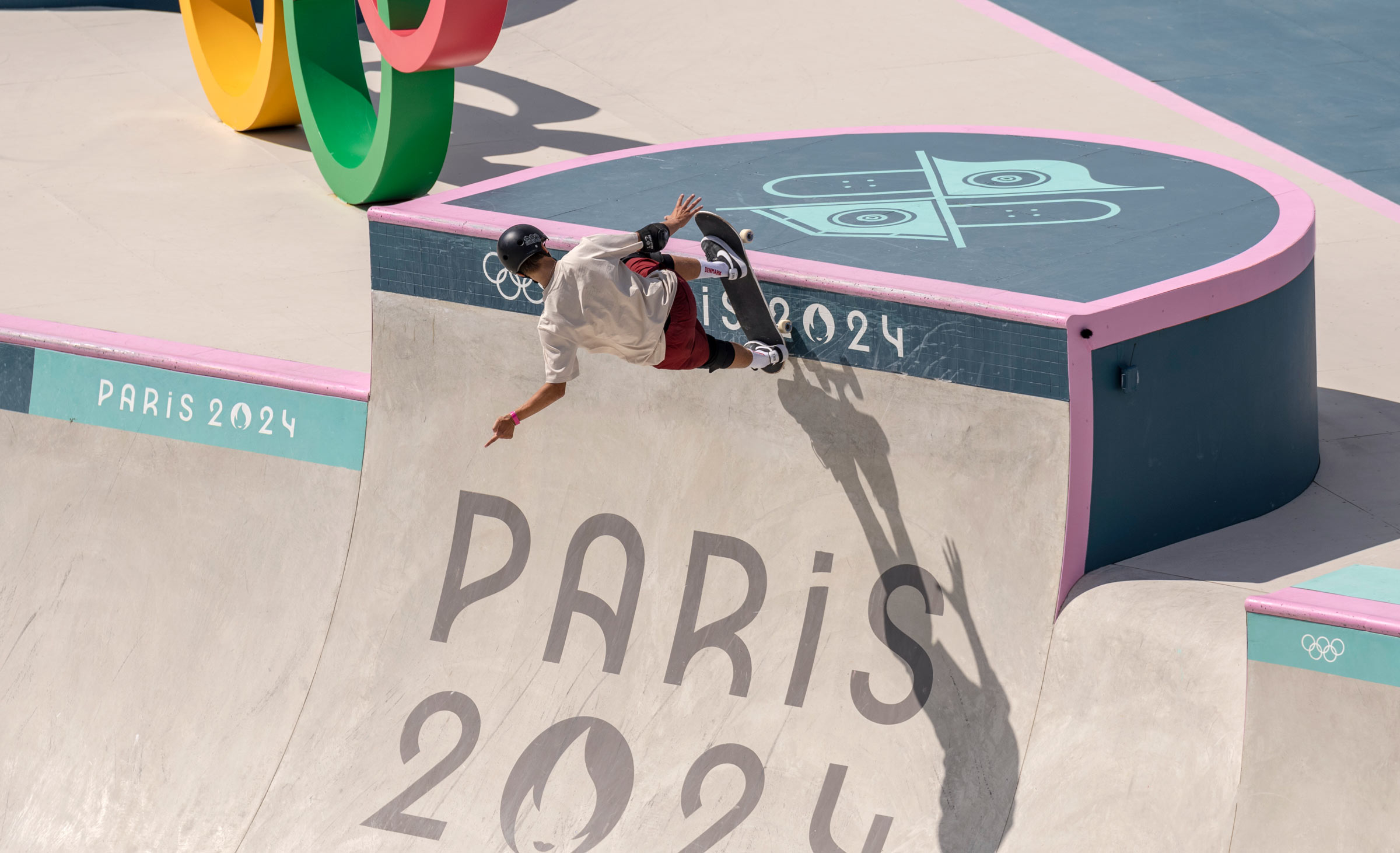 A skateboarder wearing a helmet and pads rides the vertical wall of a skatepark at the Paris 2024 Olympic Games. The curved concrete surface is marked with “PARIS 2024” and Olympic symbols.