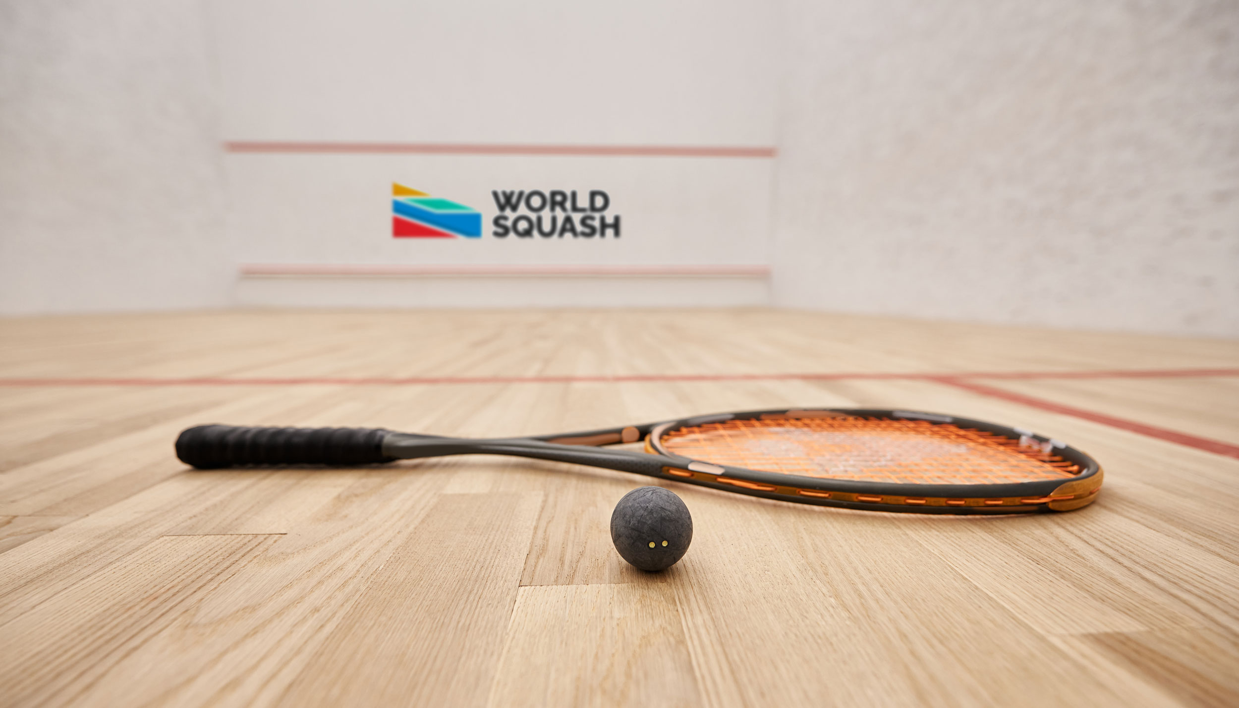 A squash ball and racquet lie on a polished wooden court. In the background, the wall features the World Squash logo with colorful geometric shapes. Red boundary lines mark the court surface inside a white-walled playing area.