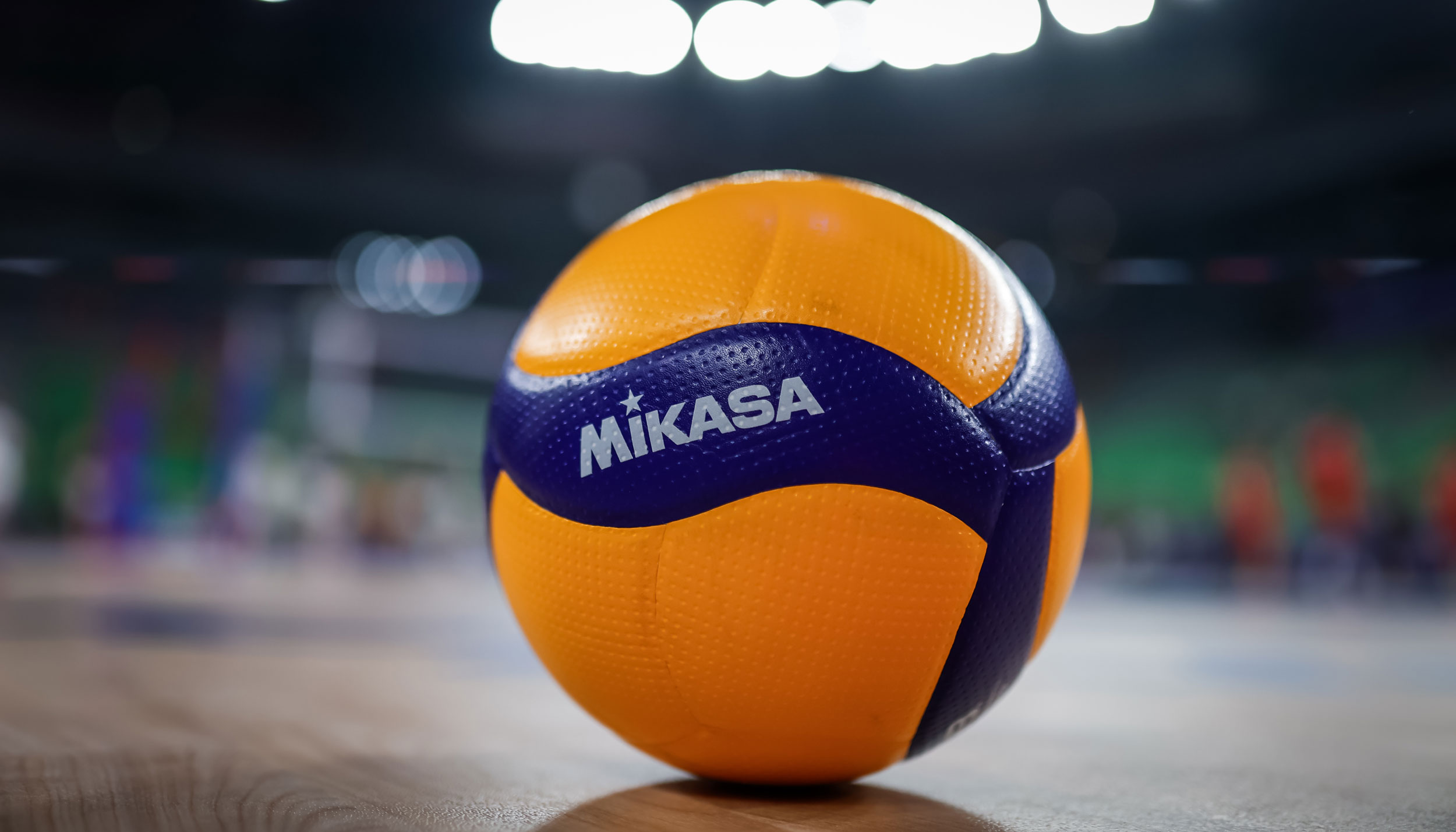 A close-up of a Mikasa volleyball resting on a polished indoor court. The ball is yellow and blue with textured panels, sharply in focus against a blurred background of a brightly lit arena.
