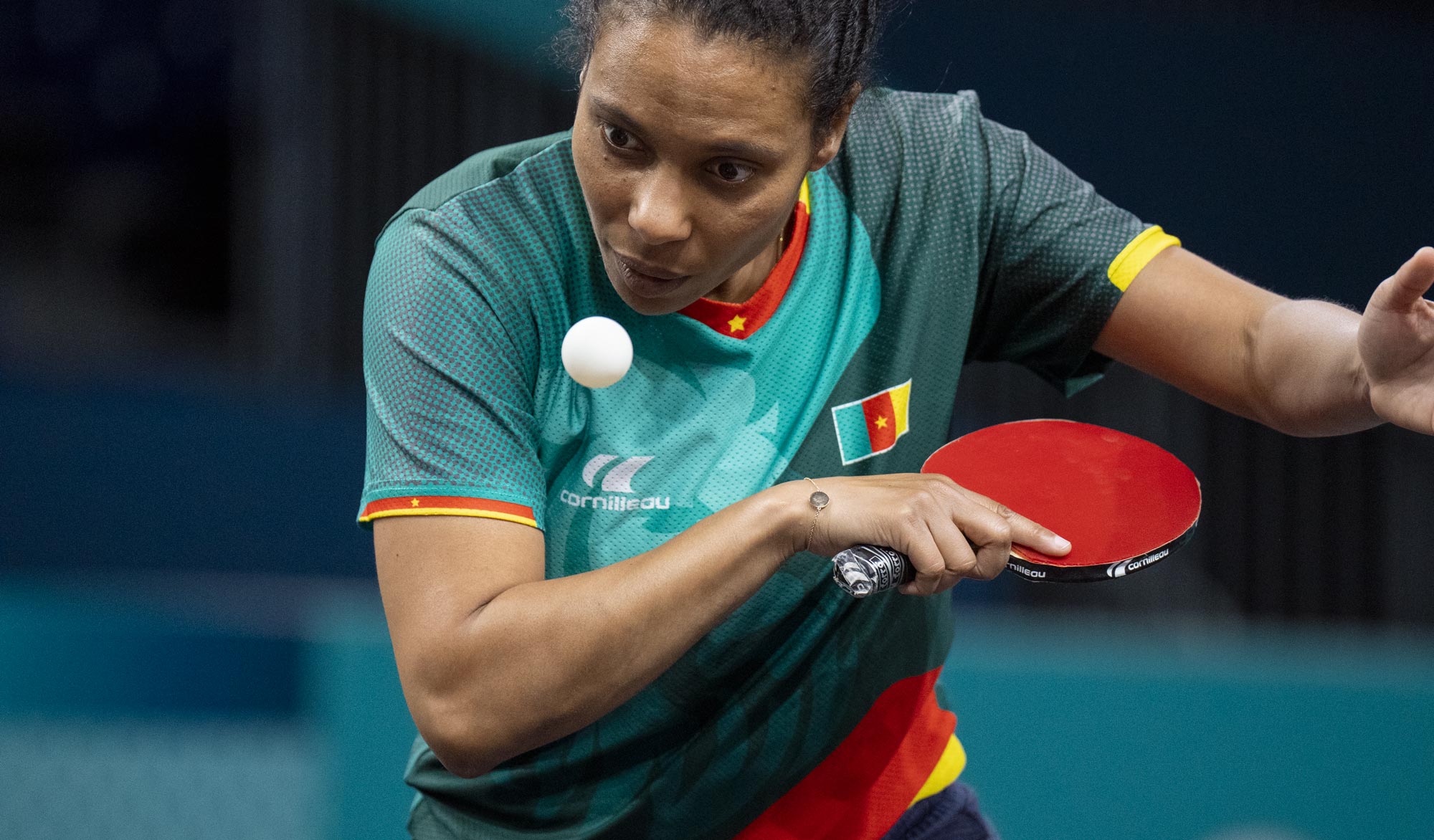 A focused female table tennis player from Cameroon prepares to serve, eyes locked on the ball mid-air. She wears a green jersey with red, yellow, and gold trim, featuring the Cameroonian flag. She grips a red paddle in her right hand and wears a silver bracelet