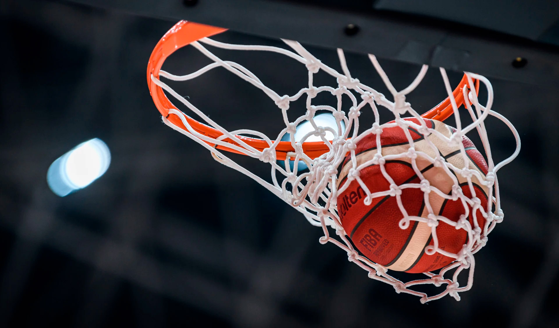 A basketball caught in mid-air as it passes cleanly through the net of an orange hoop. The ball, marked “Molten” and “FIBA,” is tightly framed from below, with the white net stretching around it and arena lights blurred in the dark background.