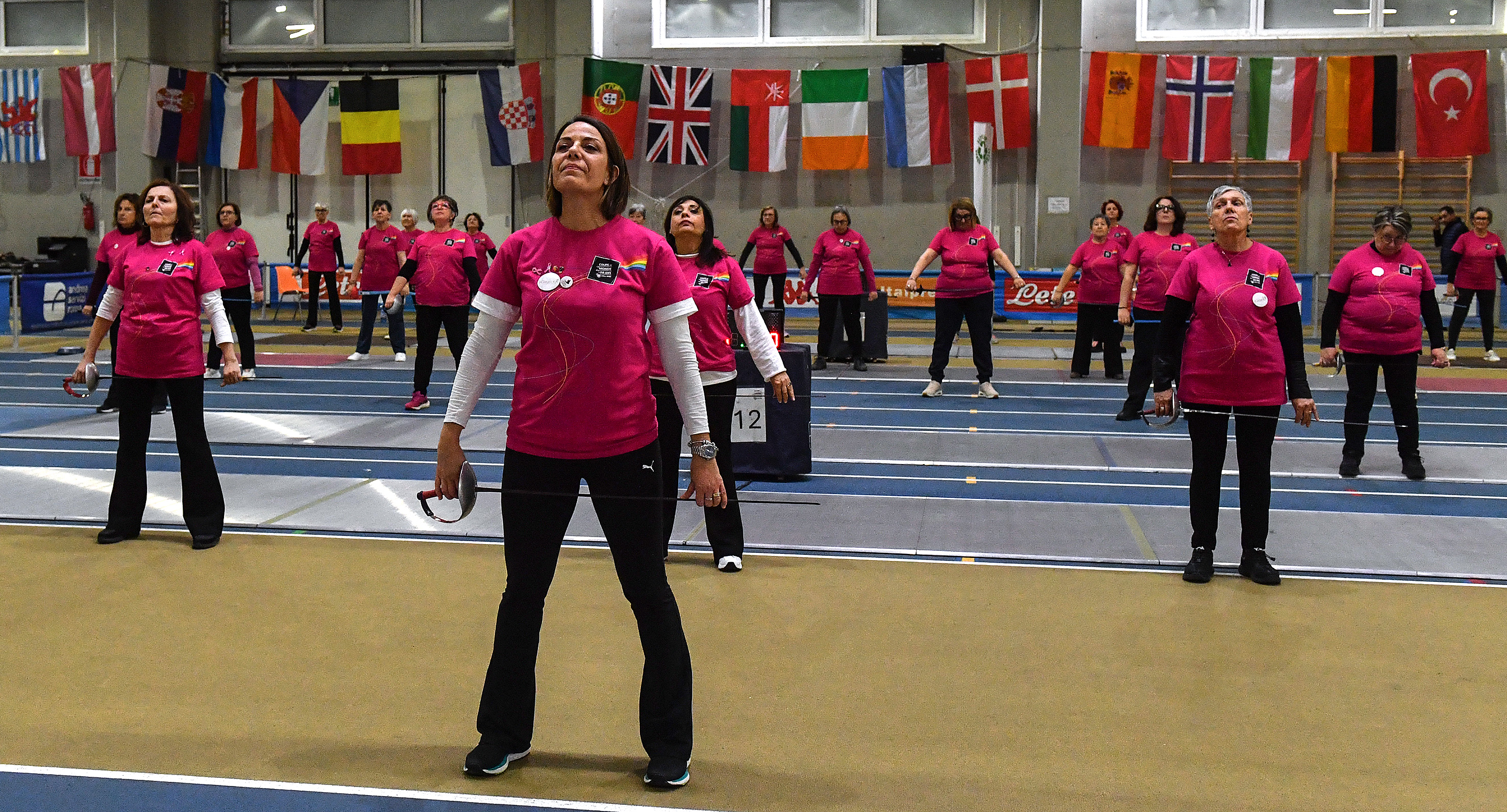 A diverse group of women stand evenly spaced in rows on a fencing piste, holding fencing foils by their sides. They wear matching bright pink shirts and participate in a group training session. A wall behind them displays numerous international flags.