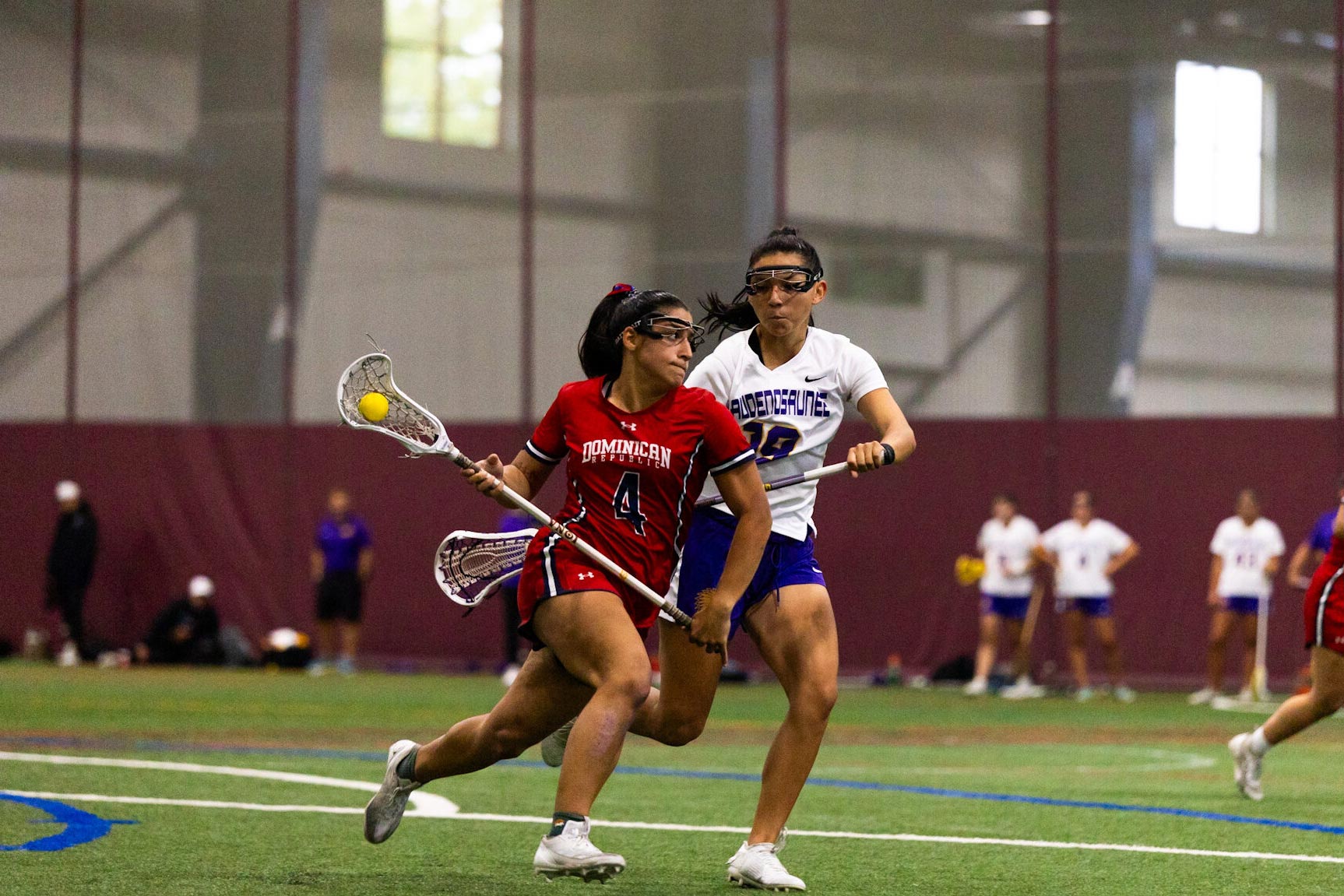 Two women’s lacrosse players compete indoors, one in red from Dominican Republic cradling the ball, closely defended by a player in white and blue.
