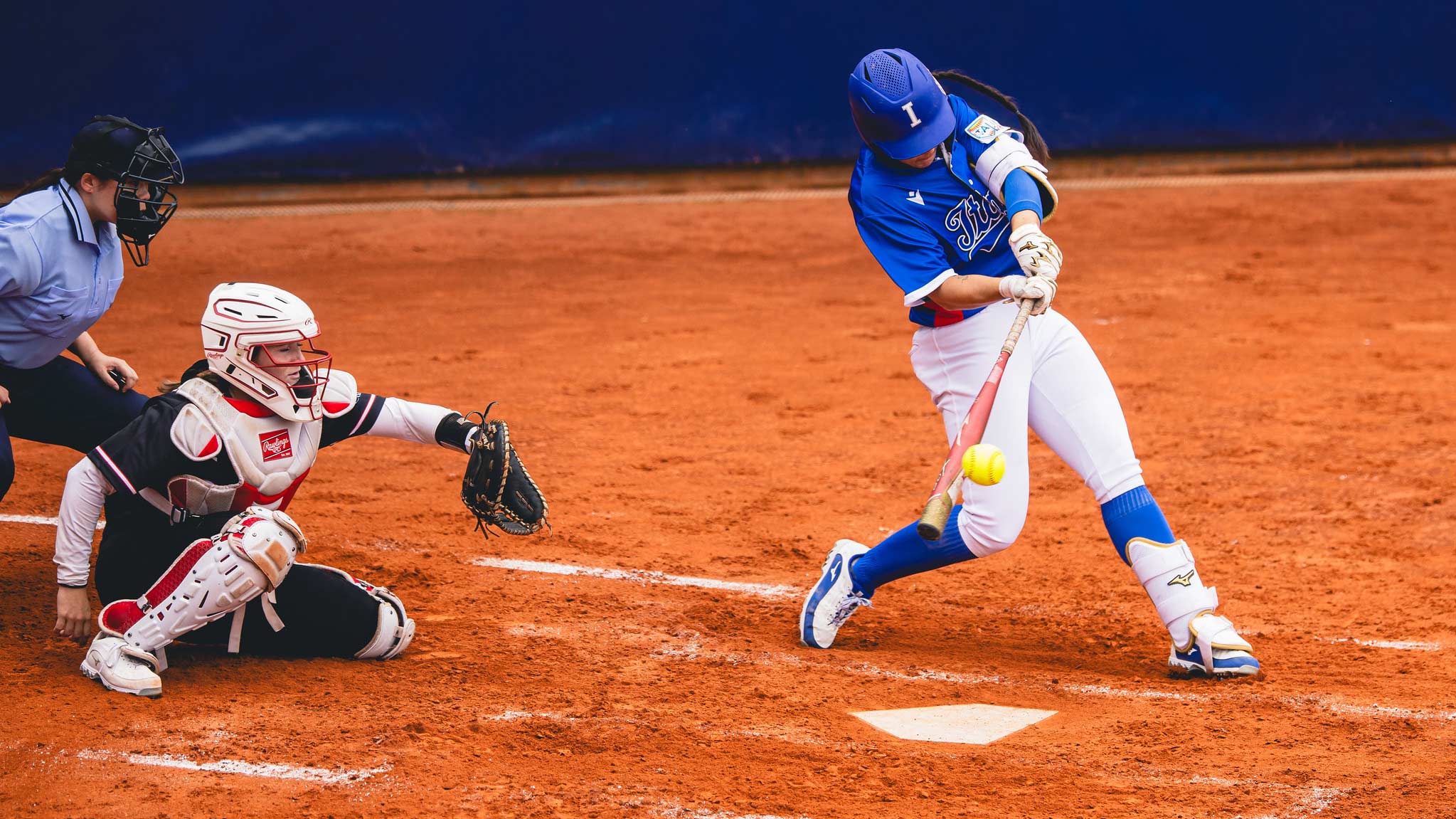 A softball player in a blue uniform swings and makes contact with the ball at home plate, while the catcher and umpire watch closely from behind on a clay infield.