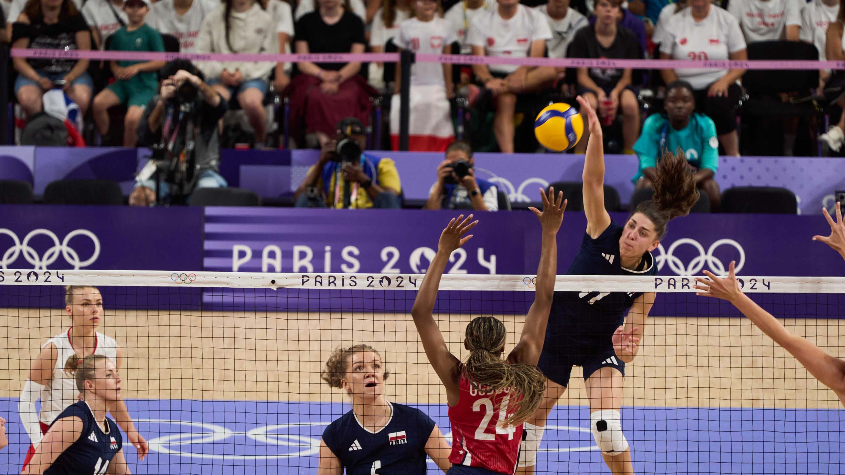 A volleyball player leaps above the net to spike the ball during a Paris 2024 Olympic match, while opposing players prepare to block and teammates watch the play unfold.