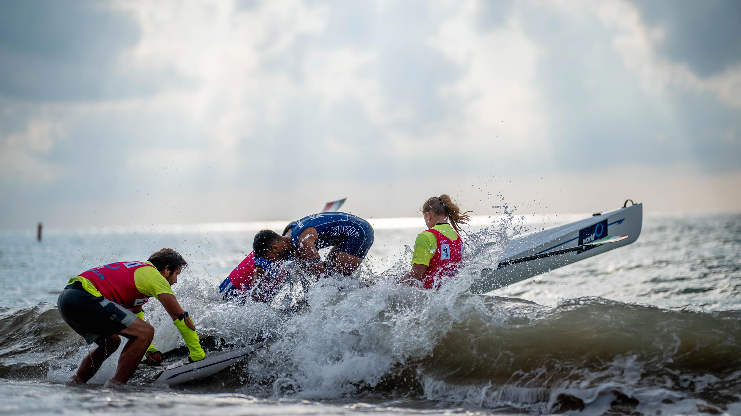 Rowers and event staff battle through breaking waves while launching a rowing boat from a beach into the sea.