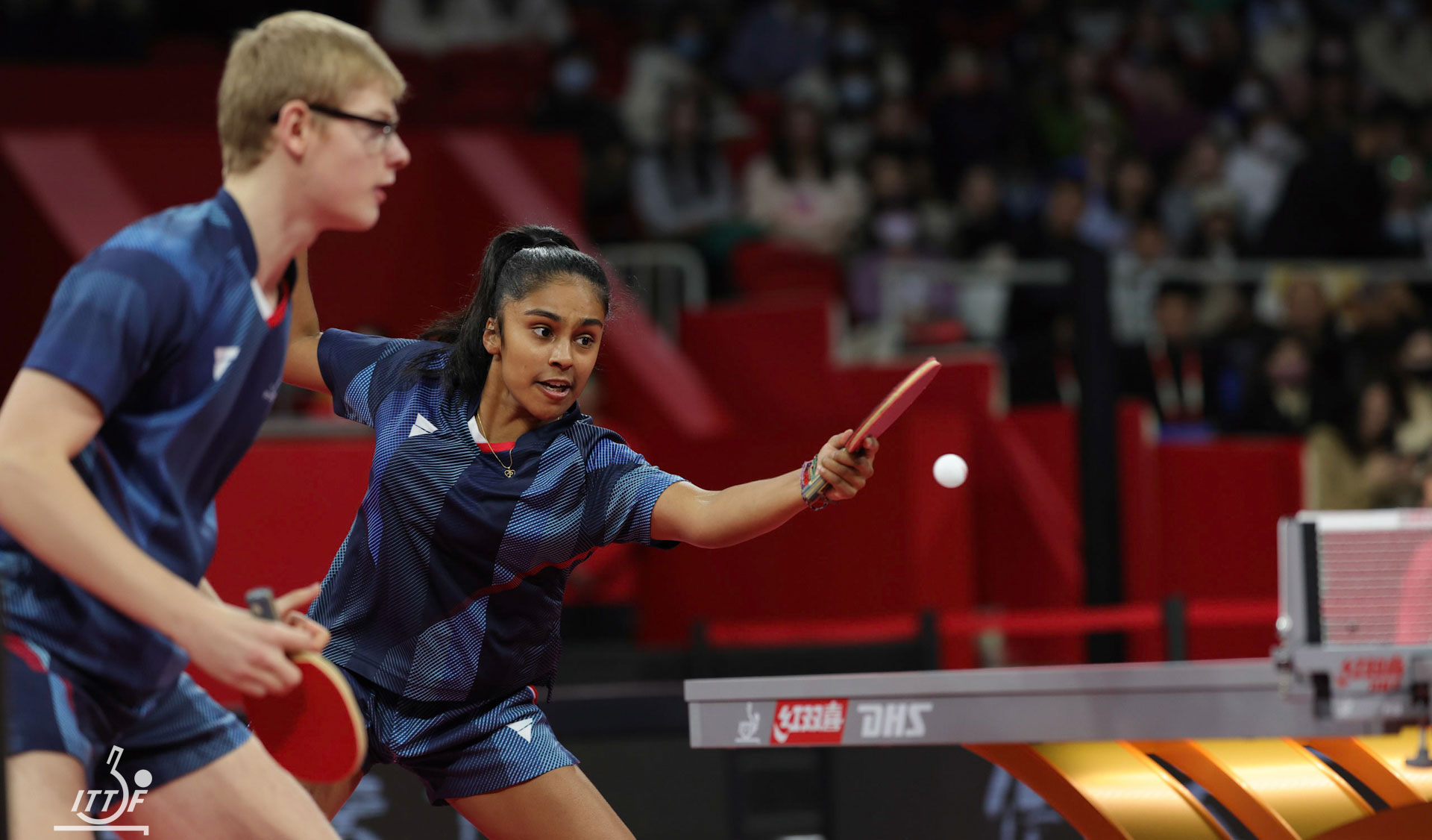 A mixed doubles table tennis pair in blue uniforms compete during a match, with the female player reaching forward to return the ball.