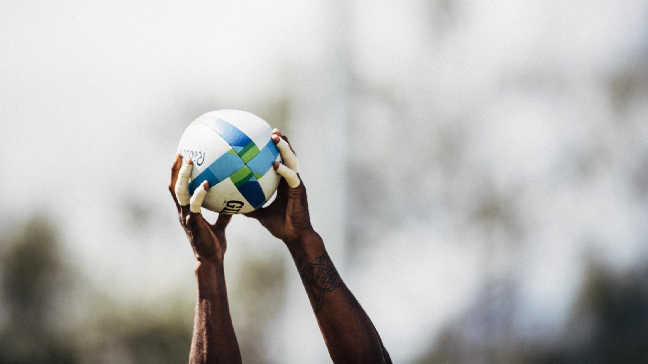 Alt text: Close-up of a rugby player’s tattooed arms holding a rugby ball overhead, preparing for a lineout throw, against a blurred stadium background.