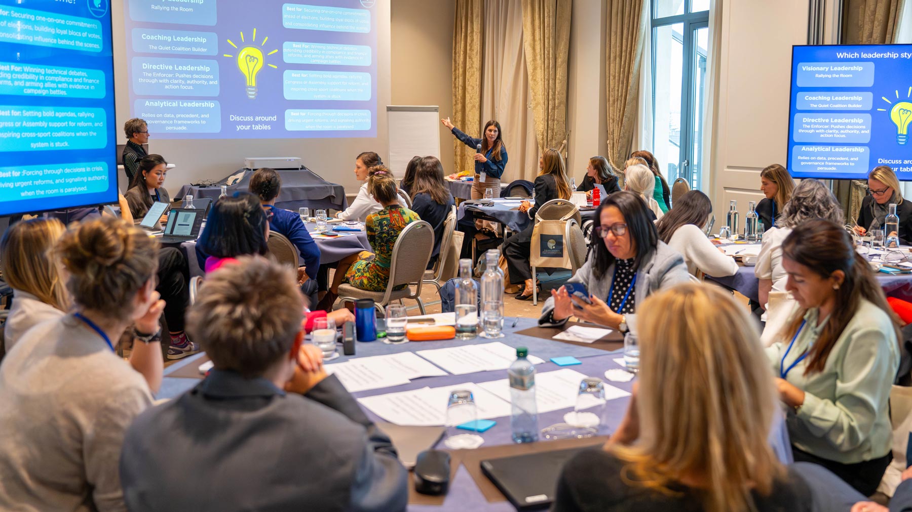 Women participating in a leadership workshop discuss activities around tables while a facilitator presents different leadership styles at the front of the room.
