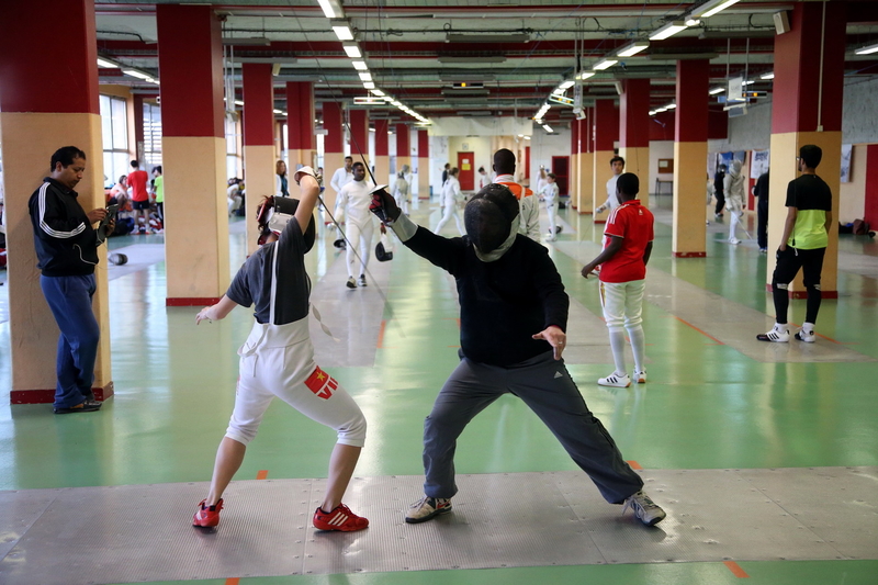 Two fencers practise together during a training session in a large indoor fencing hall, with other athletes training in the background.