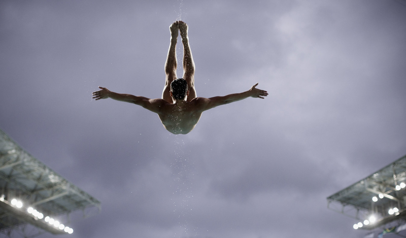 A male diver, seen from behind mid-air against a cloudy sky, stretches his arms wide and holds his feet together above his head during a dive.