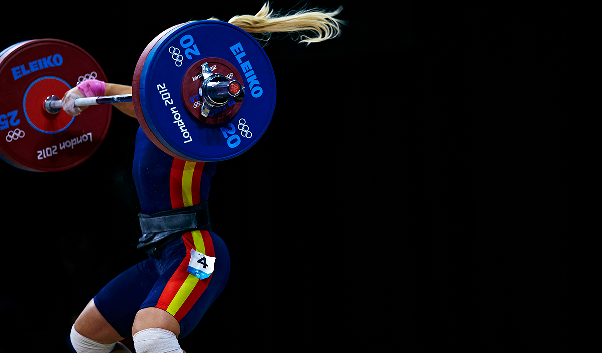 A female weightlifter with long blonde hair lifts a heavy barbell during the London 2012 Olympics. She wears a navy uniform with red and yellow vertical stripes and grips the bar with chalked hands. The colored weight plates are marked with "ELEIKO" and Olympic rings, set against a black background.