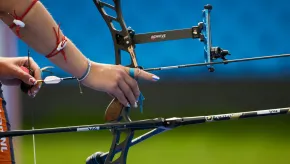 Close-up of a female archer’s hands preparing to shoot. The athlete grips a modern recurve bow with a nocked arrow and precision sight. Her nails are painted, and her arms are decorated with red cords and bracelets. The background is a blurred blue and green field.