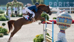 A French equestrian rider in blue and white uniform guides a chestnut horse mid-jump over a decorated obstacle during a show jumping event. The colorful jump includes a blue and gold orb with classical motifs. A blurred “PARIS 2024” sign and flower arrangements appear in the background.