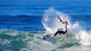 A female surfer carves across the face of a wave, sending a high spray of water into the air. She is wearing a white competition jersey and black wetsuit bottoms, balancing skillfully on her board against the deep blue ocean backdrop.