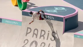 A skateboarder wearing a helmet and pads rides the vertical wall of a skatepark at the Paris 2024 Olympic Games. The curved concrete surface is marked with “PARIS 2024” and Olympic symbols.