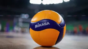 A close-up of a Mikasa volleyball resting on a polished indoor court. The ball is yellow and blue with textured panels, sharply in focus against a blurred background of a brightly lit arena.