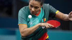 A focused female table tennis player from Cameroon prepares to serve, eyes locked on the ball mid-air. She wears a green jersey with red, yellow, and gold trim, featuring the Cameroonian flag. She grips a red paddle in her right hand and wears a silver bracelet