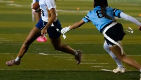 A flag football player in a white and blue jersey sprints forward holding the ball, while a defender in a light blue jersey reaches to grab one of her flag belts. The players are mid-action on a marked green field under stadium lighting.