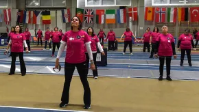 A diverse group of women stand evenly spaced in rows on a fencing piste, holding fencing foils by their sides. They wear matching bright pink shirts and participate in a group training session. A wall behind them displays numerous international flags.