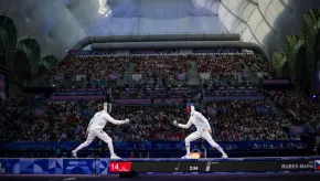 Two fencers compete on a lit piste inside a packed arena during the Paris 2024 Olympic Games, with weapons crossed mid-bout.