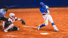 A softball player in a blue uniform swings and makes contact with the ball at home plate, while the catcher and umpire watch closely from behind on a clay infield.