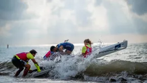 Rowers and event staff battle through breaking waves while launching a rowing boat from a beach into the sea.