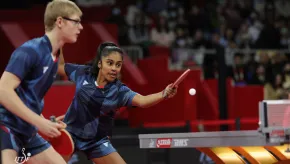A mixed doubles table tennis pair in blue uniforms compete during a match, with the female player reaching forward to return the ball.