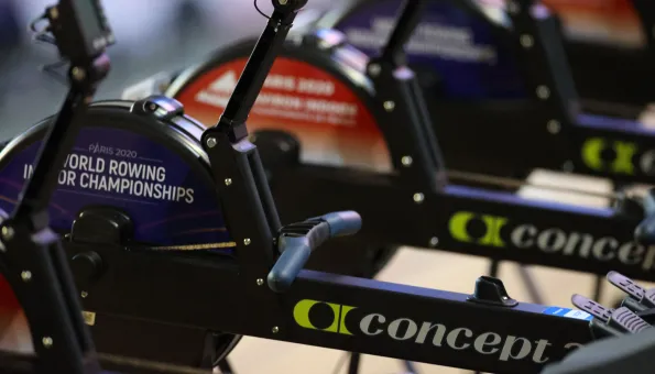 A close-up view of several indoor rowing machines lined up side by side. The machines are branded "Concept2", and one features a visible round panel labeled "Paris 2020 World Rowing Indoor Championships". The camera focuses on the flywheel and handle area, emphasizing the competitive, professional setup.