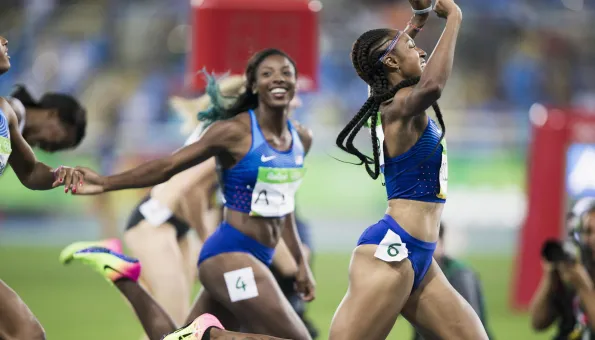 A jubilant moment at a track event as a sprinter with braided hair, wearing a blue USA uniform and bib number 6, celebrates a win with her arms raised. Her teammate, also in blue and smiling widely, reaches out to clasp her hand in mid-stride. The scene is energetic, with other runners and a blurred stadium crowd in the background.