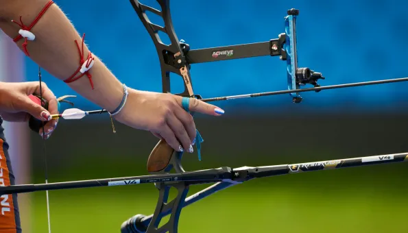Close-up of a female archer’s hands preparing to shoot. The athlete grips a modern recurve bow with a nocked arrow and precision sight. Her nails are painted, and her arms are decorated with red cords and bracelets. The background is a blurred blue and green field.