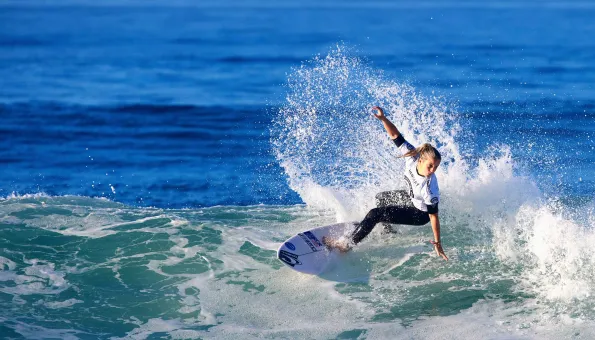 A female surfer carves across the face of a wave, sending a high spray of water into the air. She is wearing a white competition jersey and black wetsuit bottoms, balancing skillfully on her board against the deep blue ocean backdrop.