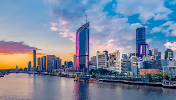 A vibrant city skyline of Brisbane, Australia at sunset. Skyscrapers reflect hues of pink and orange in the evening light, with the Brisbane River flowing calmly in the foreground. A colorful sky with scattered clouds completes the scenic urban view.