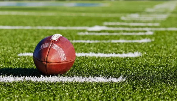 A close-up of an American football resting on a green artificial turf field, with white yard lines stretching diagonally into the distance. The ball’s laces and textured surface are clearly visible under bright daylight.