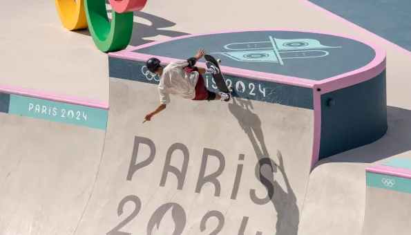 A skateboarder wearing a helmet and pads rides the vertical wall of a skatepark at the Paris 2024 Olympic Games. The curved concrete surface is marked with “PARIS 2024” and Olympic symbols.