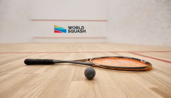 A squash ball and racquet lie on a polished wooden court. In the background, the wall features the World Squash logo with colorful geometric shapes. Red boundary lines mark the court surface inside a white-walled playing area.
