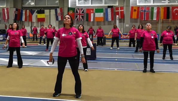 A diverse group of women stand evenly spaced in rows on a fencing piste, holding fencing foils by their sides. They wear matching bright pink shirts and participate in a group training session. A wall behind them displays numerous international flags.