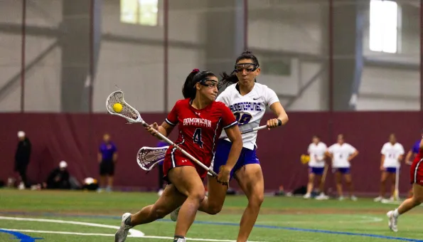 Two women’s lacrosse players compete indoors, one in red from Dominican Republic cradling the ball, closely defended by a player in white and blue.