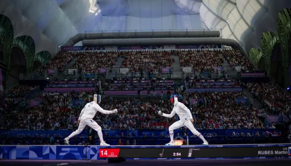 Two fencers compete on a lit piste inside a packed arena during the Paris 2024 Olympic Games, with weapons crossed mid-bout.