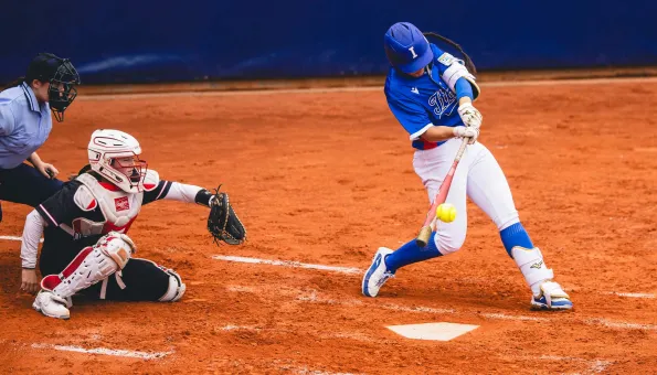 A softball player in a blue uniform swings and makes contact with the ball at home plate, while the catcher and umpire watch closely from behind on a clay infield.