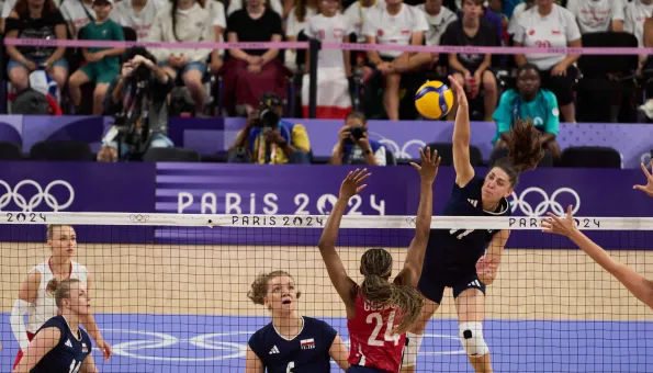 A volleyball player leaps above the net to spike the ball during a Paris 2024 Olympic match, while opposing players prepare to block and teammates watch the play unfold.
