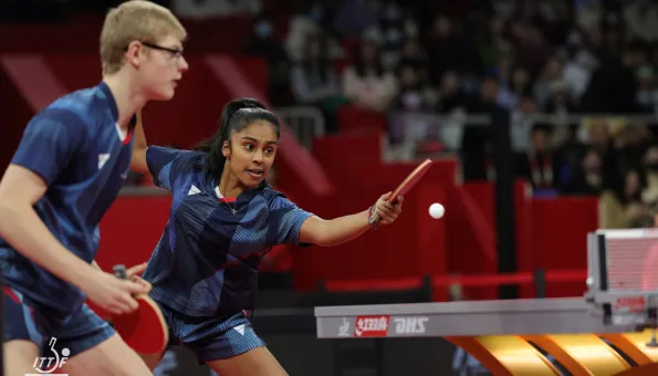A mixed doubles table tennis pair in blue uniforms compete during a match, with the female player reaching forward to return the ball.