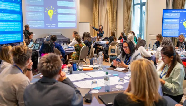Women participating in a leadership workshop discuss activities around tables while a facilitator presents different leadership styles at the front of the room.