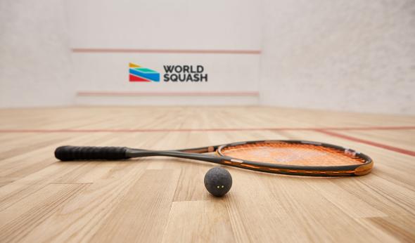 A squash ball and racquet lie on a polished wooden court. In the background, the wall features the World Squash logo with colorful geometric shapes. Red boundary lines mark the court surface inside a white-walled playing area.