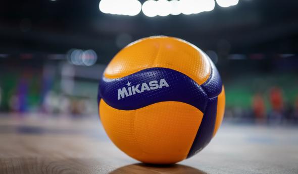 A close-up of a Mikasa volleyball resting on a polished indoor court. The ball is yellow and blue with textured panels, sharply in focus against a blurred background of a brightly lit arena.