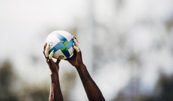 Alt text: Close-up of a rugby player’s tattooed arms holding a rugby ball overhead, preparing for a lineout throw, against a blurred stadium background.